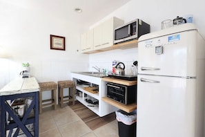 Cozinha compacta equipada com micro-ondas, forno e frigorífico. Possui dois taburetes e uma mesa pequena com chaleira.