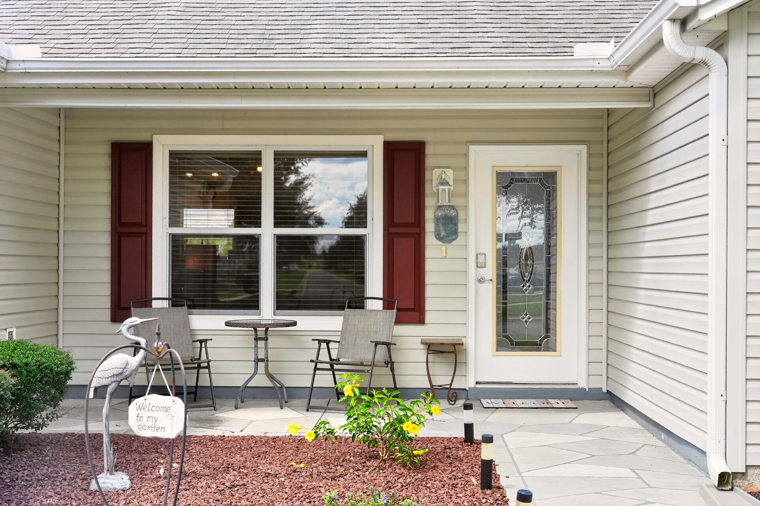 Ein Haus mit überdachter Veranda, zwei Stühlen, einem Tisch, einem Willkommensschild, einer dekorativen Vogelplastik und einem kleinen Garten.