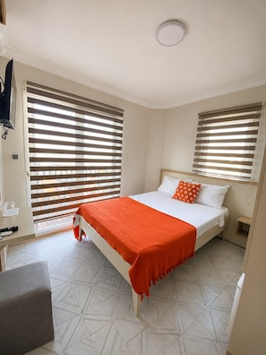 Un dormitorio con cama, dos mesitas de noche, televisor montado en la pared y ventana con persianas.