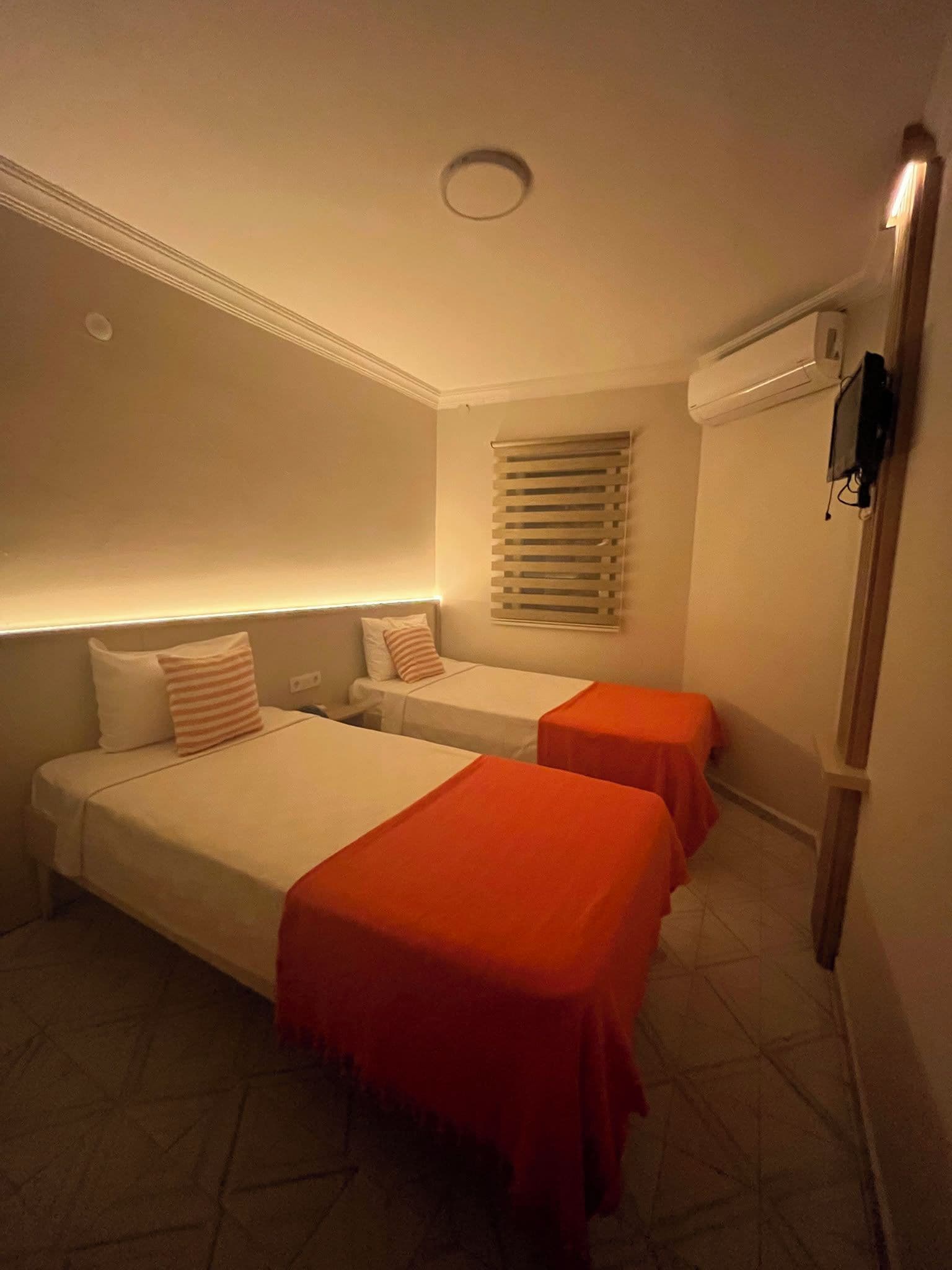 Habitación de hotel con dos camas, una ventana con persianas, aire acondicionado instalado en la pared y un televisor de pantalla plana.