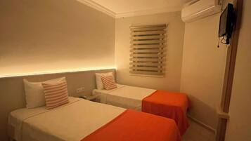 Habitación de hotel con dos camas, una ventana con persianas, aire acondicionado instalado en la pared y un televisor de pantalla plana.