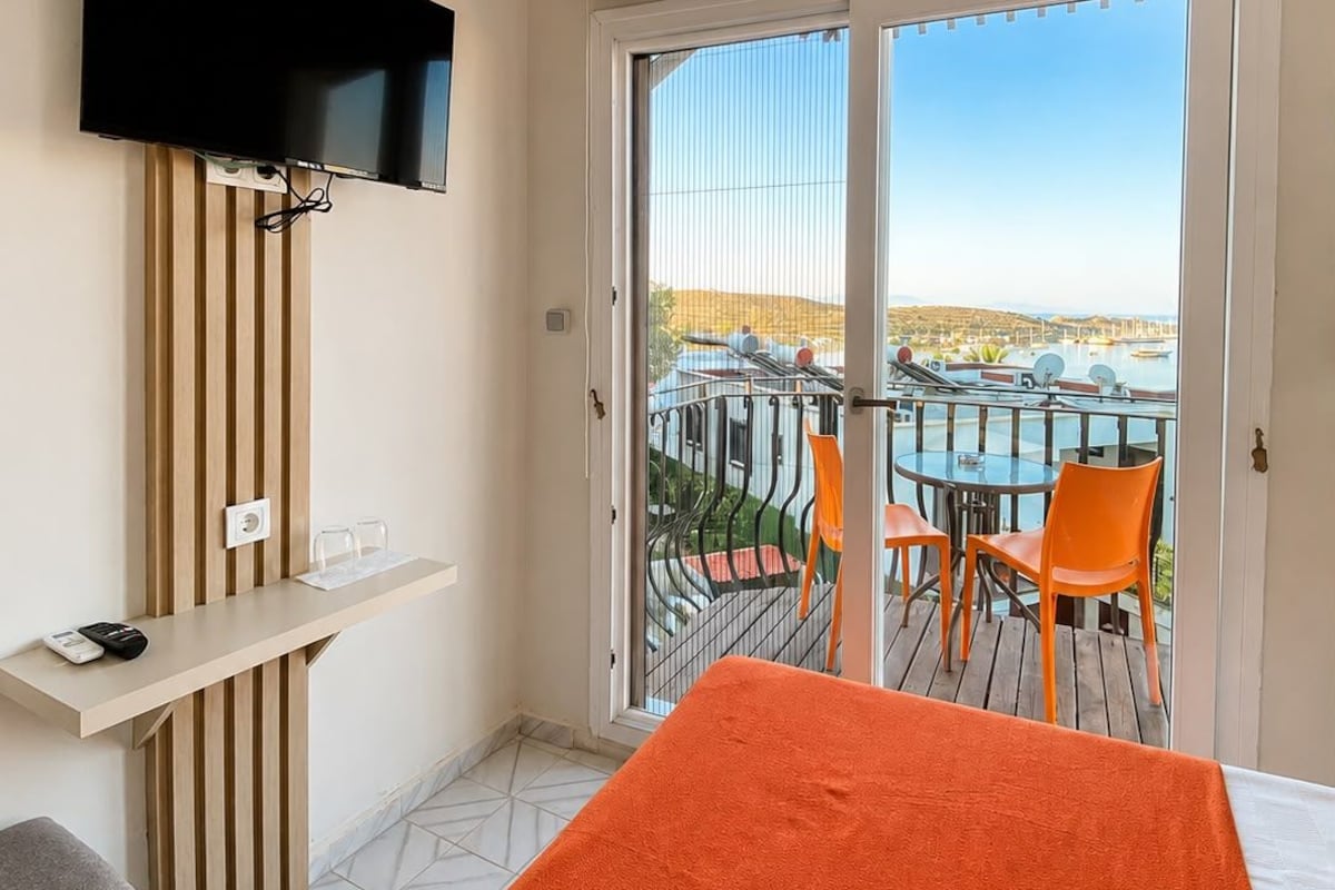 A hotel room with a bed, a TV mounted on a wooden panel, a small table with a remote control, and a balcony with a table and chairs.
