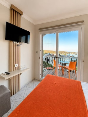 Habitación de hotel con cama, televisor montado en un panel de madera, una mesita con un control remoto, y un balcón con mesa y sillas.