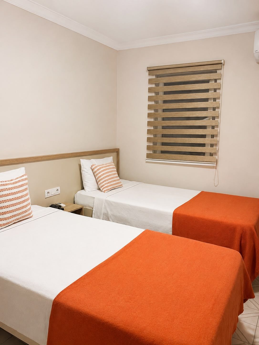 A hotel room with two beds, each with a white bedspread and orange throw pillows. There is a window with wooden blinds, and a small wooden nightstand with a lamp.