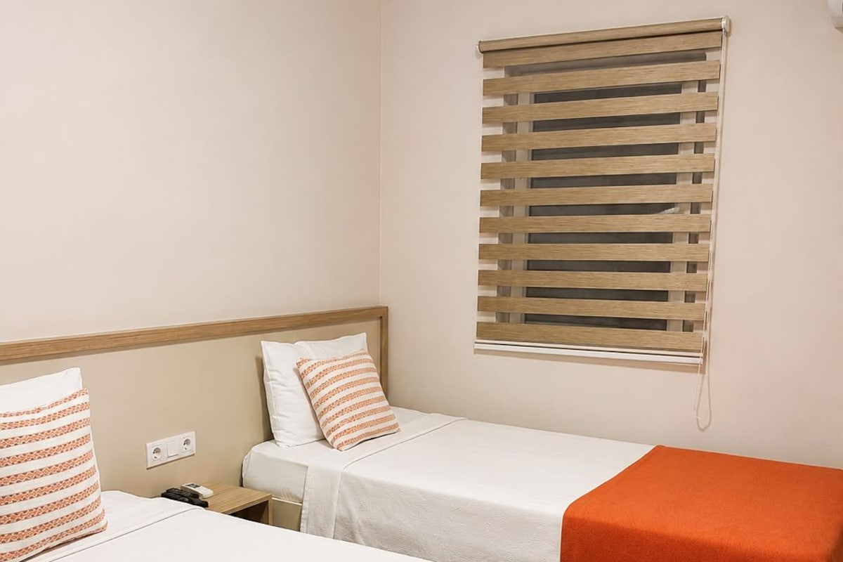 Habitación de hotel con dos camas, cada una con cubrecama blanco y almohadones naranjas. Se ve una ventana con persianas de madera, y una mesita de noche de madera con una lámpara.