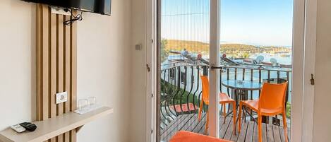 A hotel room with a bed, a TV mounted on a wooden panel, a small table with a remote control, and a balcony with a table and chairs.