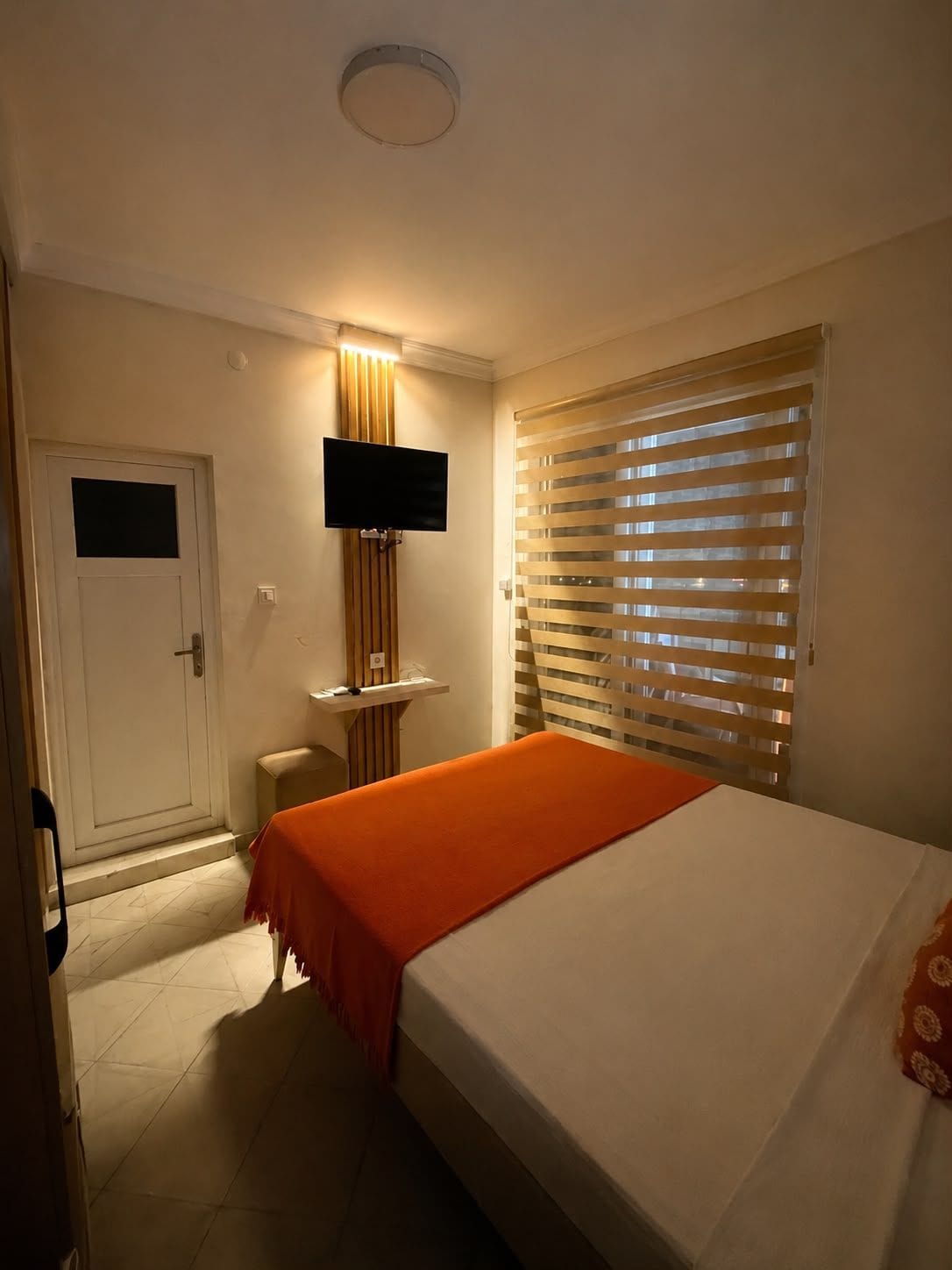 A hotel room with a bed, a television mounted on the wall, a window with blinds, and a door.