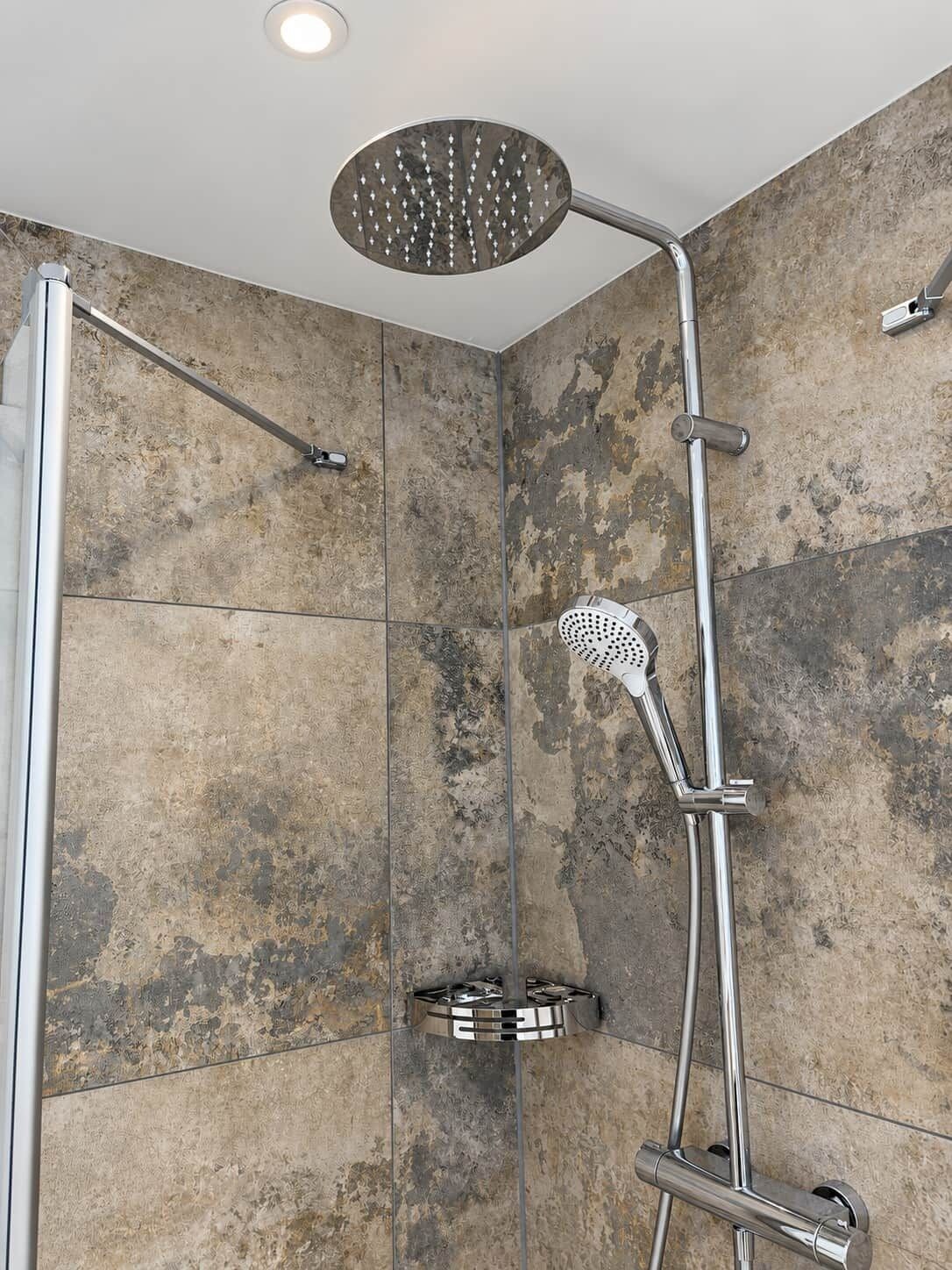 A shower with a rain showerhead and a handheld showerhead, set against a textured wall.