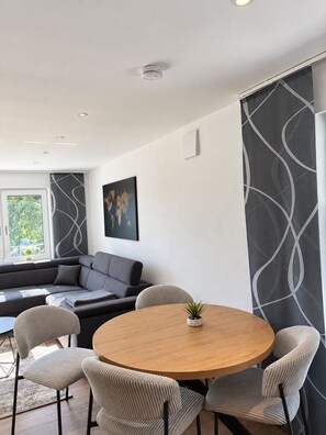 Ein stilvolles Wohnzimmer mit einem runden Holztisch, einer grauen Stoffcouch und gemusterten Vorhängen.