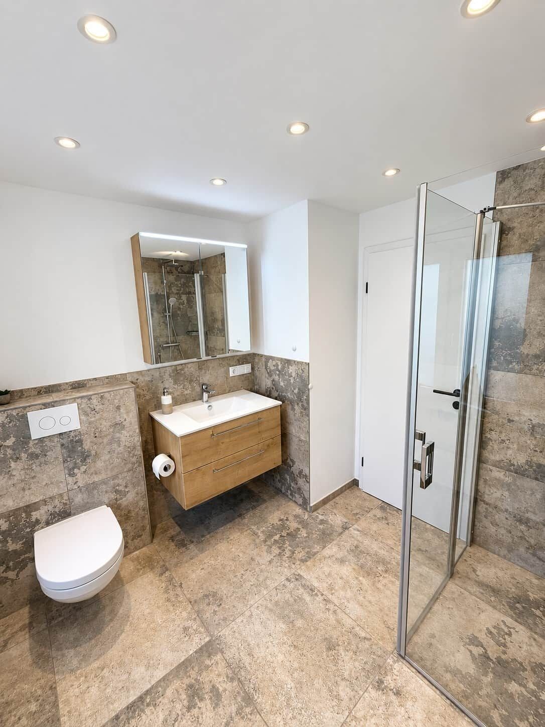 A modern bathroom with a glass shower enclosure, a white toilet, a vanity with a mirror, and a marble-tiled floor.