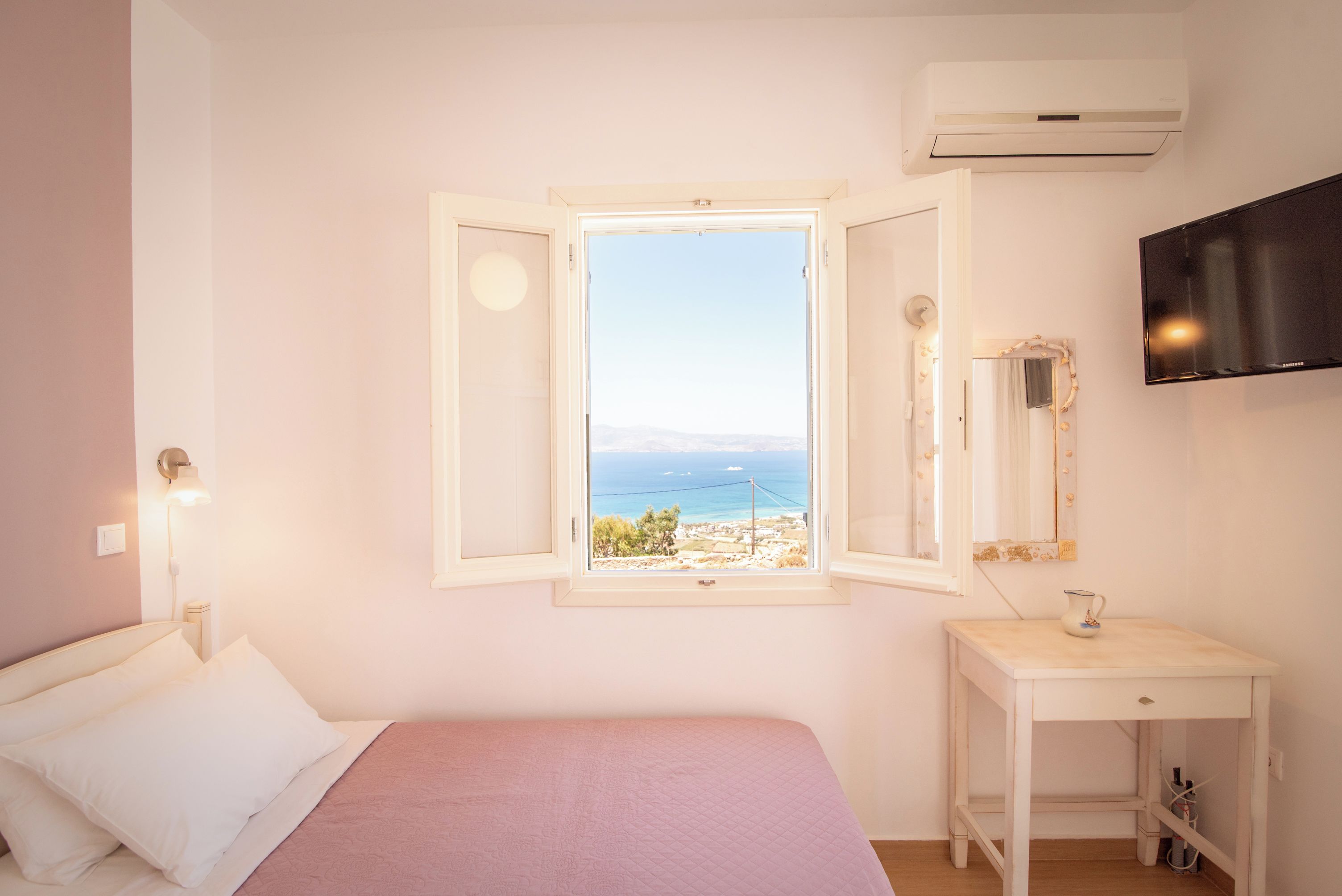 Un dormitorio con cama, mesita de noche, espejo, ventana con vista al mar y aire acondicionado.