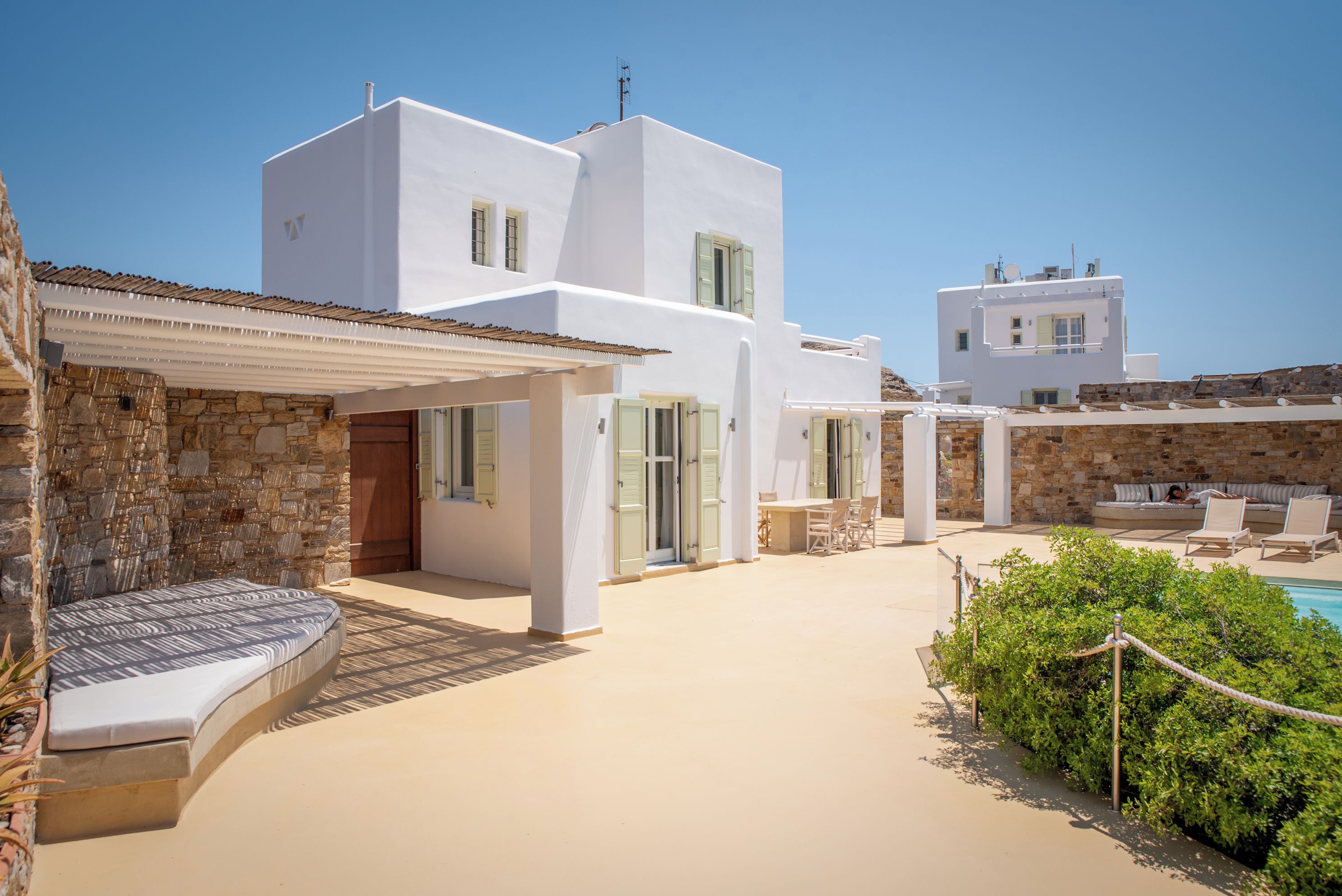 Un bâtiment moderne aux murs blancs, avec une entrée ornée d’un mur en pierre, un espace extérieur couvert avec des sièges et un coin détente au bord de la piscine, équipé de chaises longues.