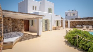 Un bâtiment moderne aux murs blancs, avec une entrée ornée d’un mur en pierre, un espace extérieur couvert avec des sièges et un coin détente au bord de la piscine, équipé de chaises longues.