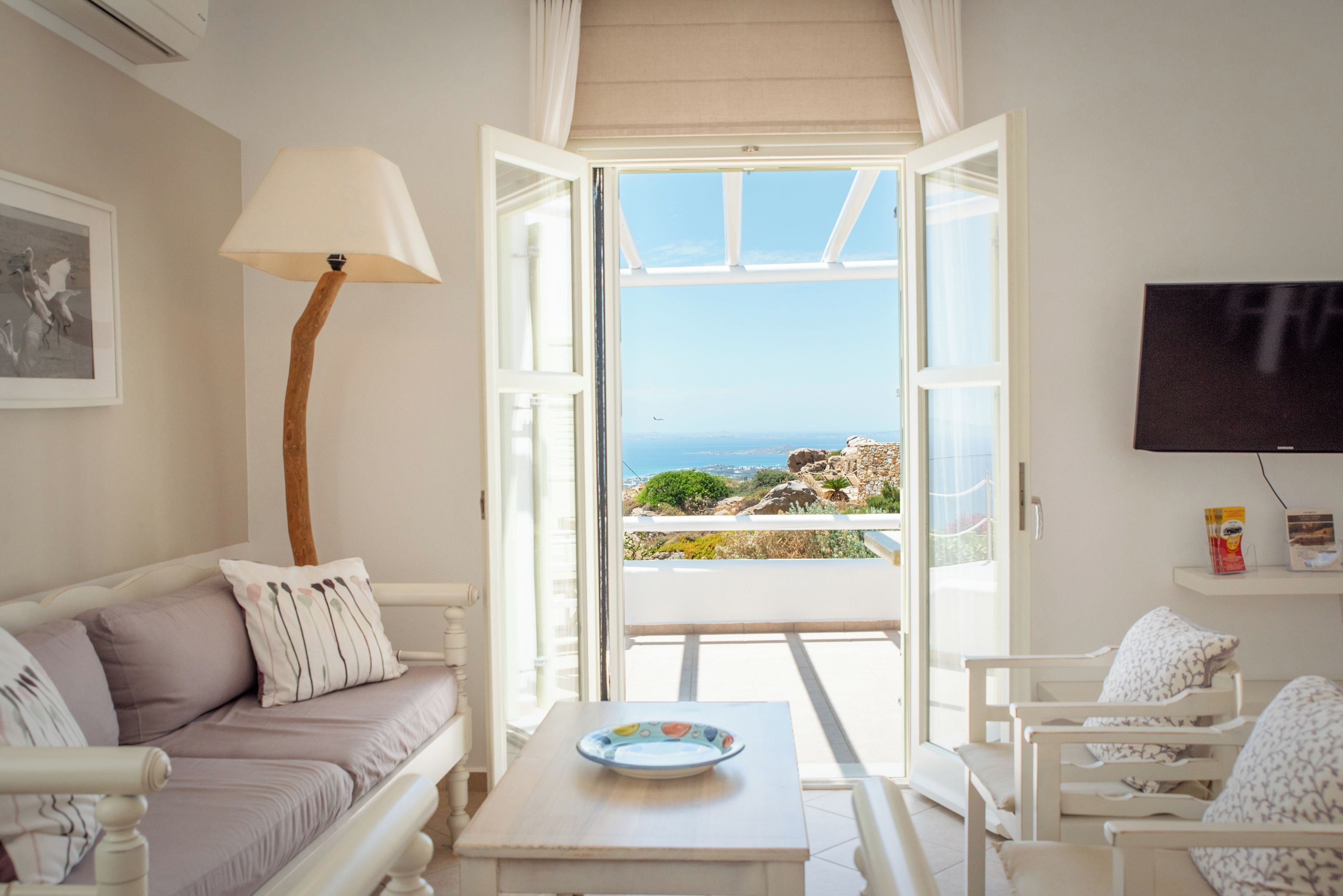 A room with a bed, a table with a plate, a lamp, a TV, and a balcony with a view of the sea.