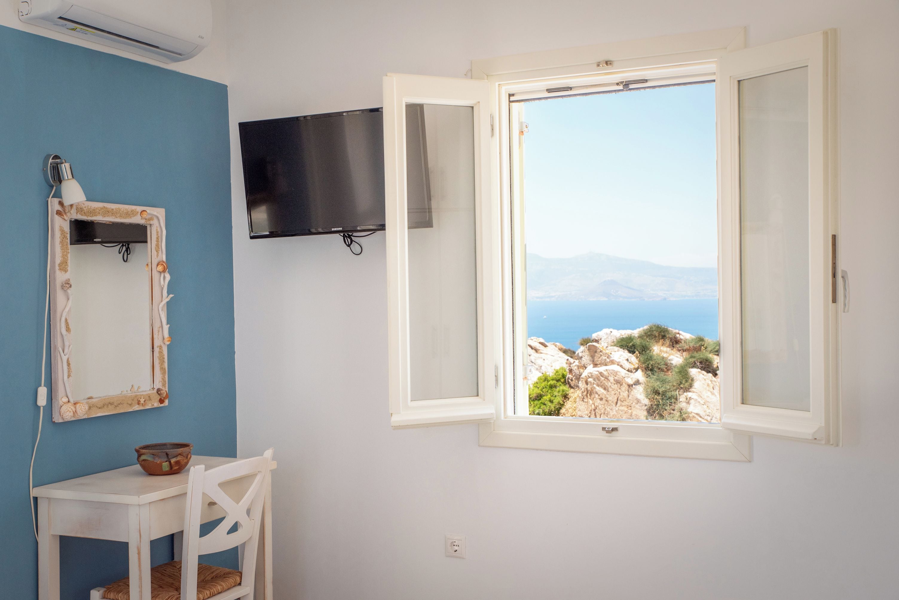 A room with a blue wall, a white desk, a chair, a television mounted on the wall, and a window with a view of a mountainous landscape.