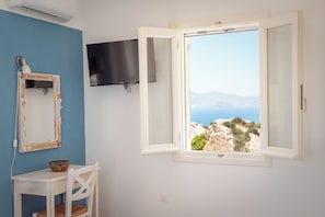 A room with a blue wall, a white desk, a chair, a television mounted on the wall, and a window with a view of a mountainous landscape.