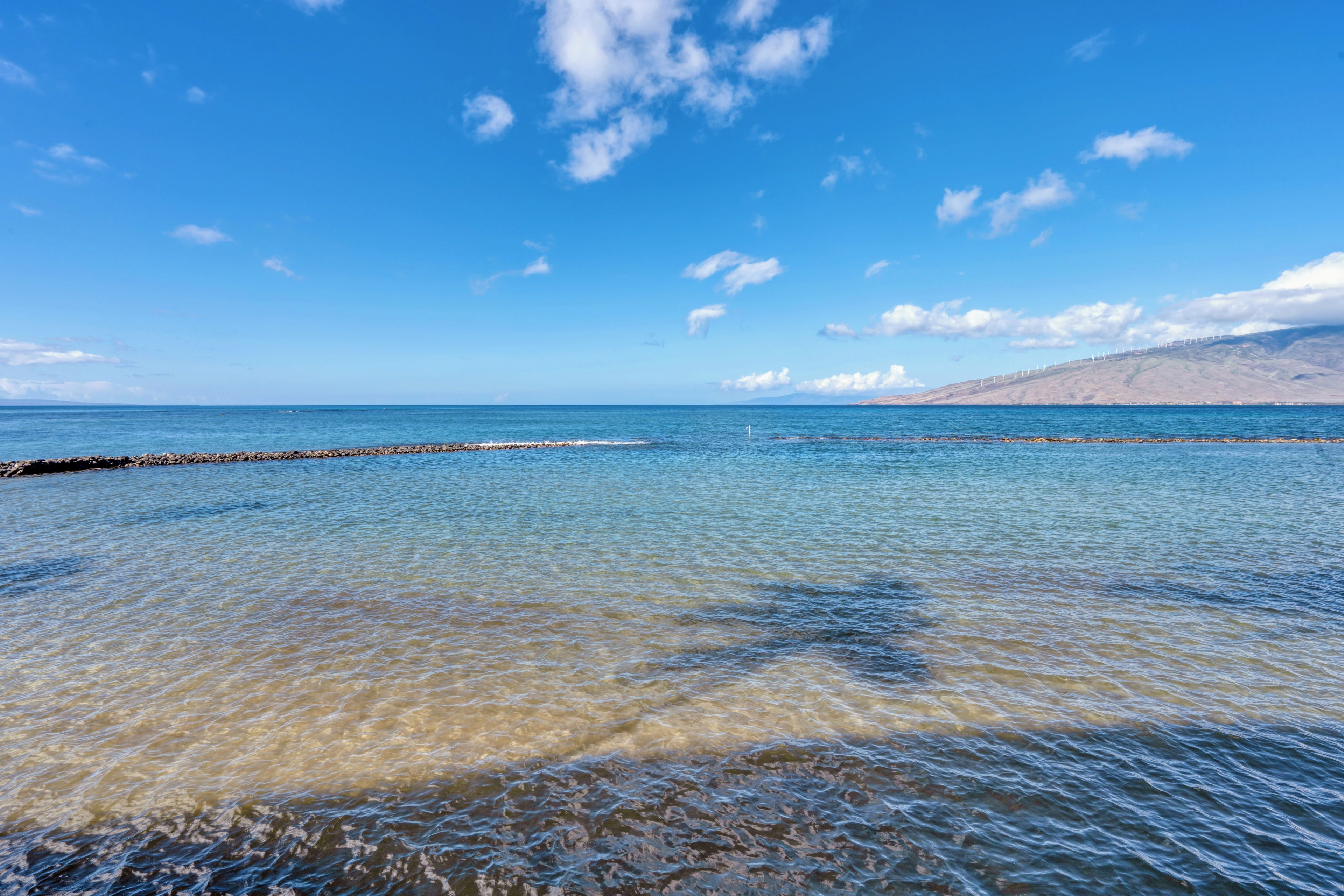 A coastal scene with clear water, a rocky breakwater, and a distant mountain range.
