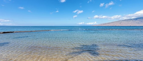 Escena costera con agua cristalina, un rompeolas rocoso y una cadena montañosa distante.