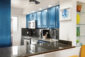 Cozinha moderna com armários azuis, bancada de granito, eletrodomésticos de aço inoxidável e prateleira de vidro com objetos decorativos.