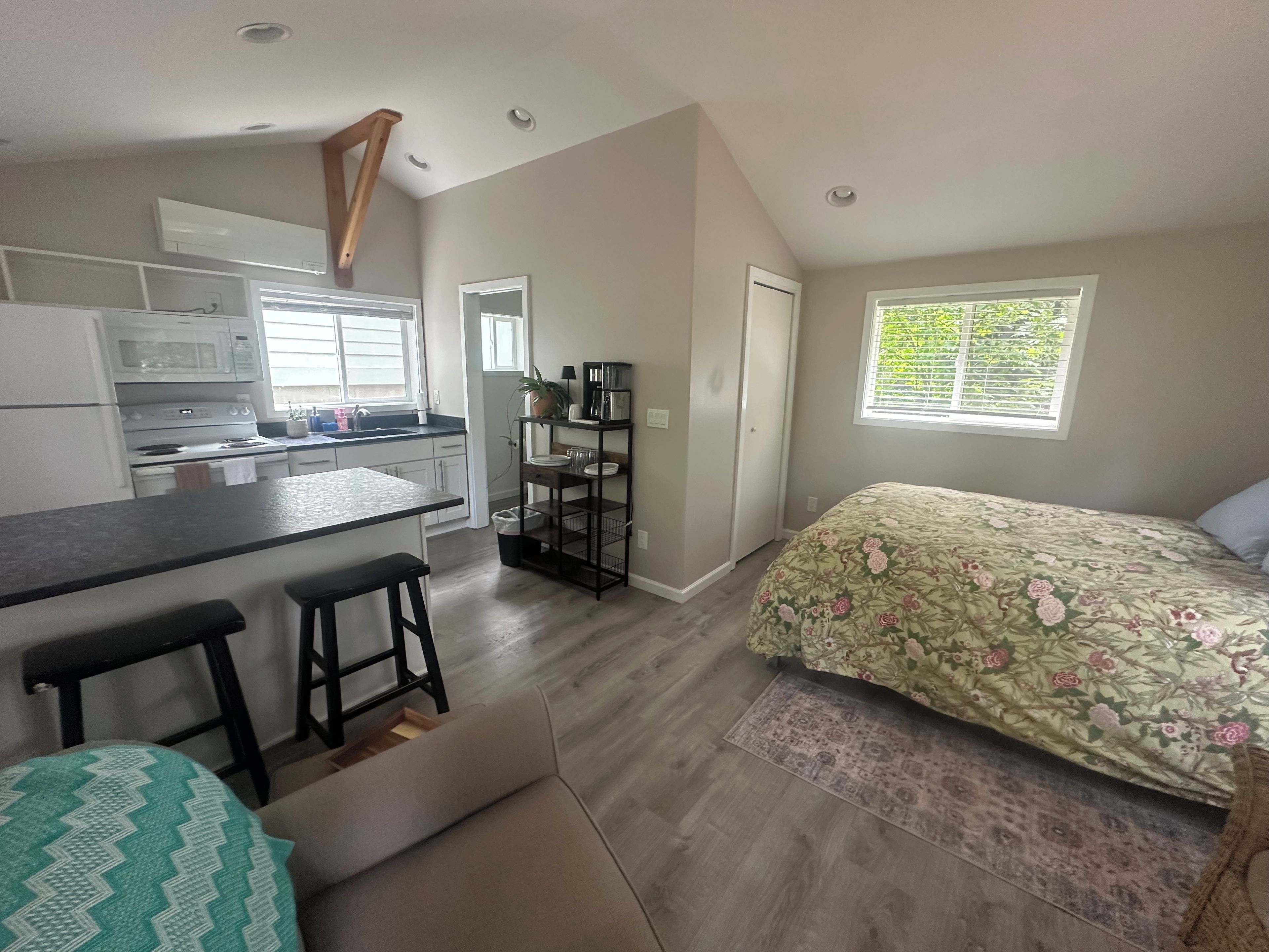 Un espacio habitable compacto con kitchenette, una cama con ropa de cama floral y una ventana con persianas.