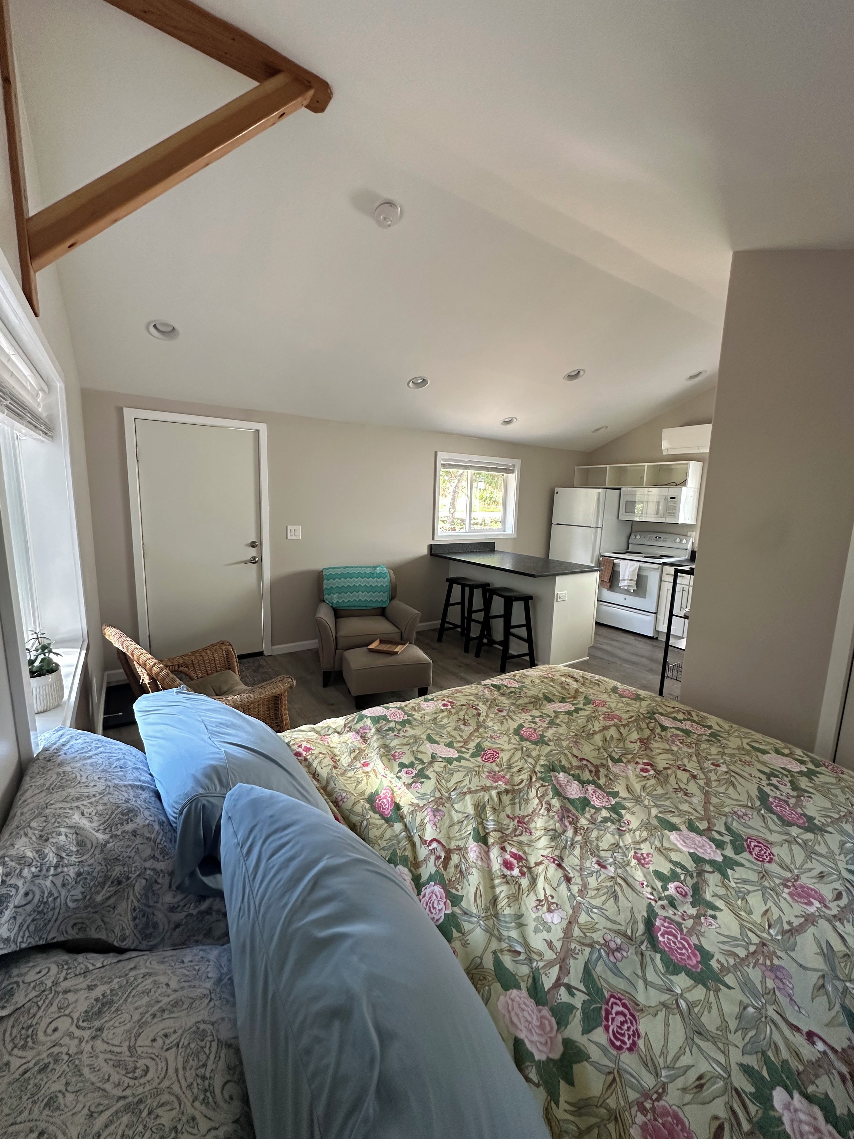 A bedroom with a bed, a chair, a small table, and a kitchenette in the background.