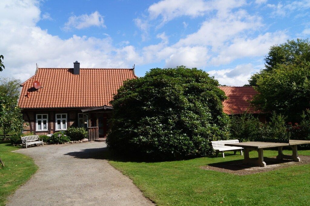 Een gebouw met een rood pannendak, een grindpad dat naar een bank leidt en een grote struik op de voorgrond.