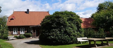 Een gebouw met een rood pannendak, een grindpad dat naar een bank leidt en een grote struik op de voorgrond.