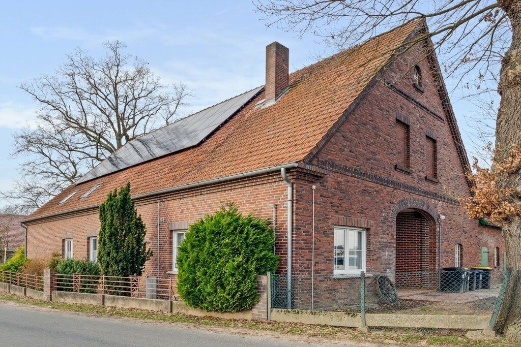A brick building with a sloped roof, a chimney, and a large bush in front.