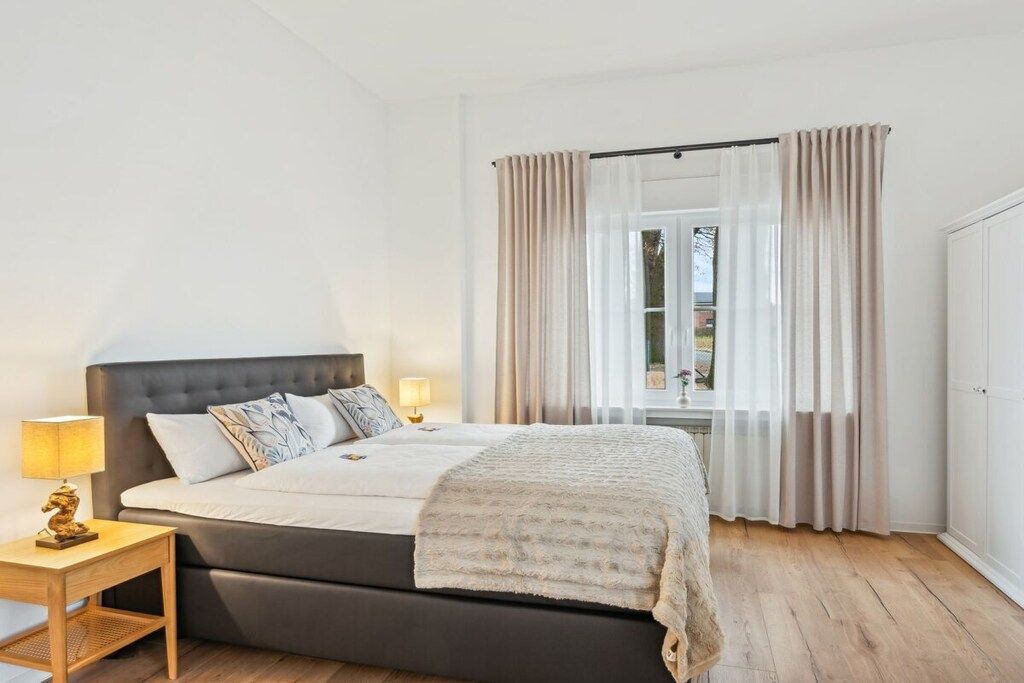 Ein Schlafzimmer mit einem großen Bett, einem Nachttisch mit Lampe und einem Fenster mit Vorhängen.