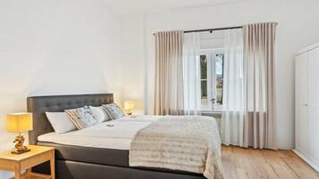 Ein Schlafzimmer mit einem großen Bett, einem Nachttisch mit Lampe und einem Fenster mit Vorhängen.