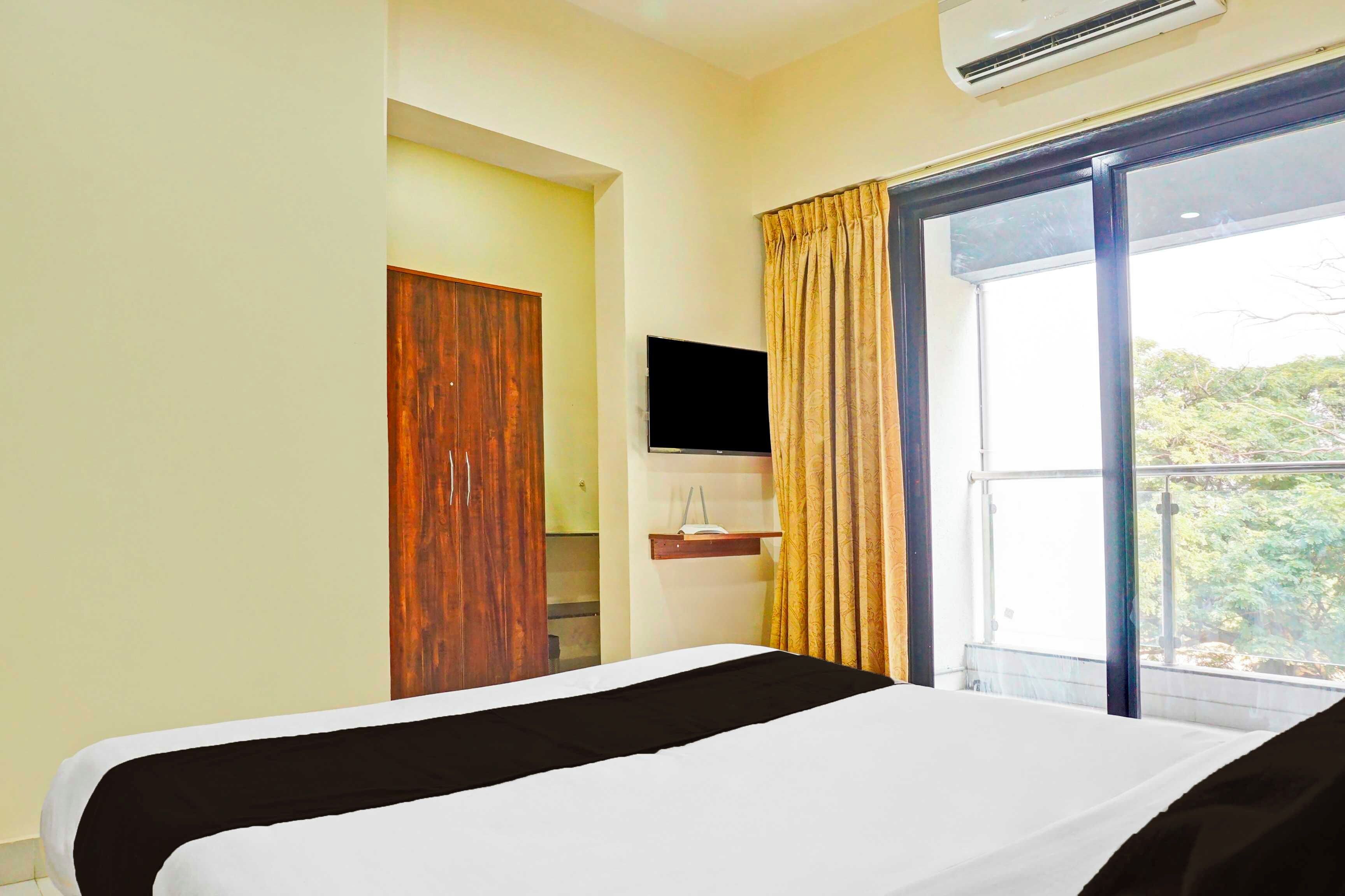 A hotel room with a bed, a television, a window with curtains, and an air conditioning unit.