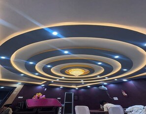 A room with a circular ceiling design, recessed lighting, and a chandelier. There are tables and chairs arranged for seating. - Hotel Laxmi Inn (Danapur)