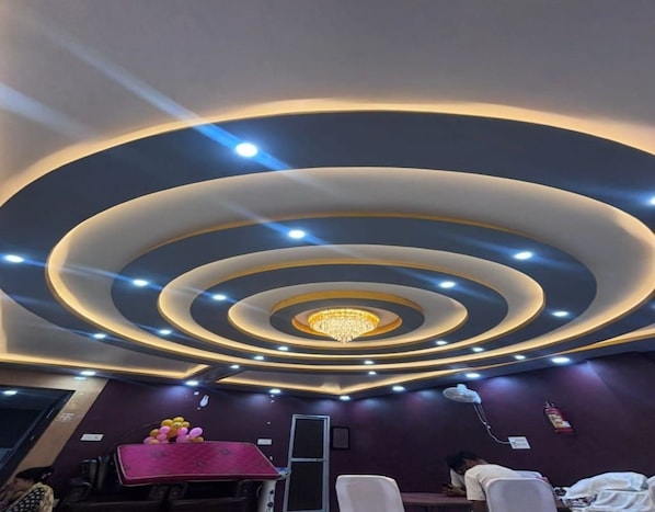 A room with a circular ceiling design, recessed lighting, and a chandelier. There are tables and chairs arranged for seating.