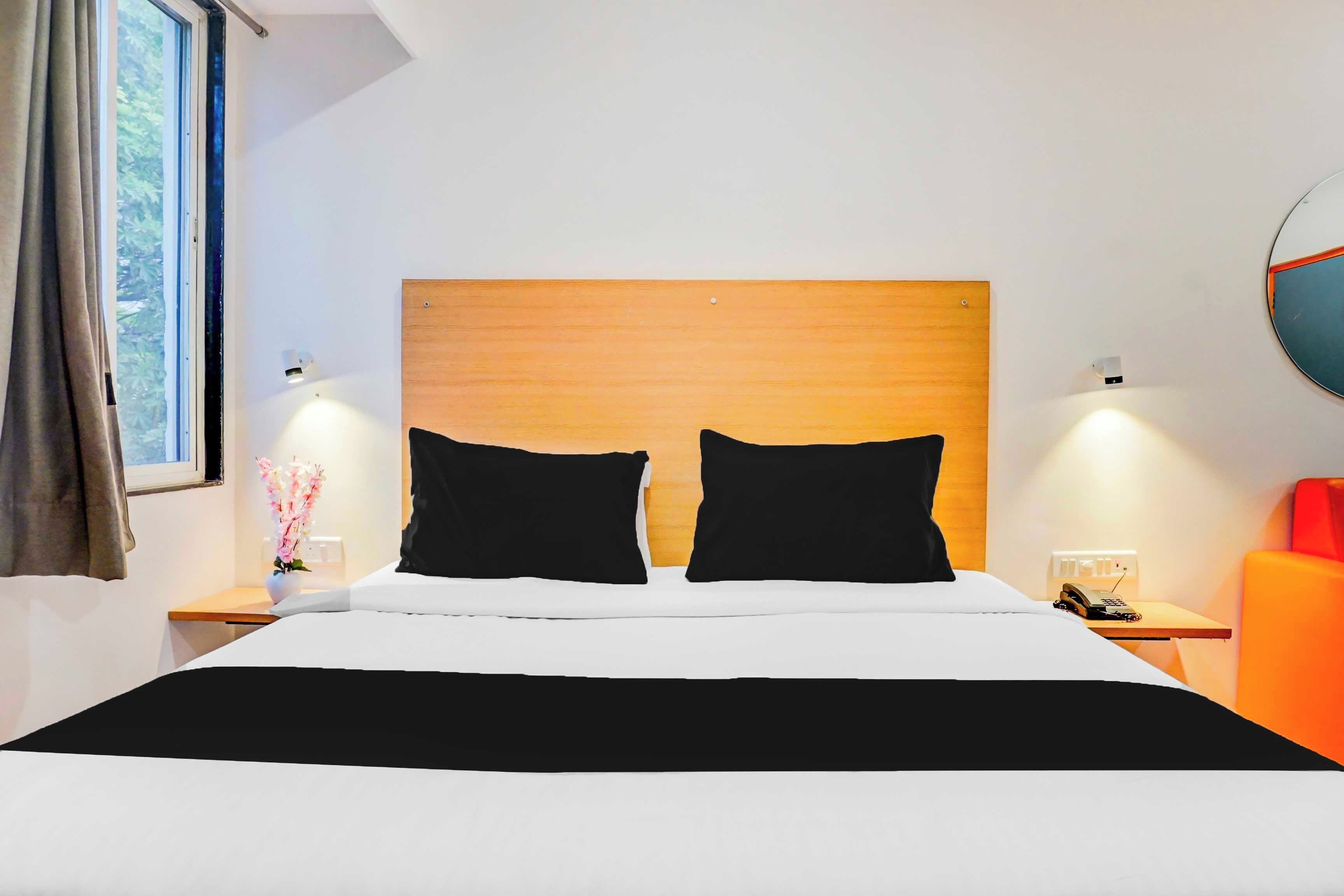 A hotel room with a wooden headboard, two black pillows, a white bedspread, a bedside table with a lamp, a telephone, and a red chair.