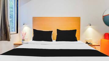 A hotel room with a wooden headboard, two black pillows, a white bedspread, a bedside table with a lamp, a telephone, and a red chair.