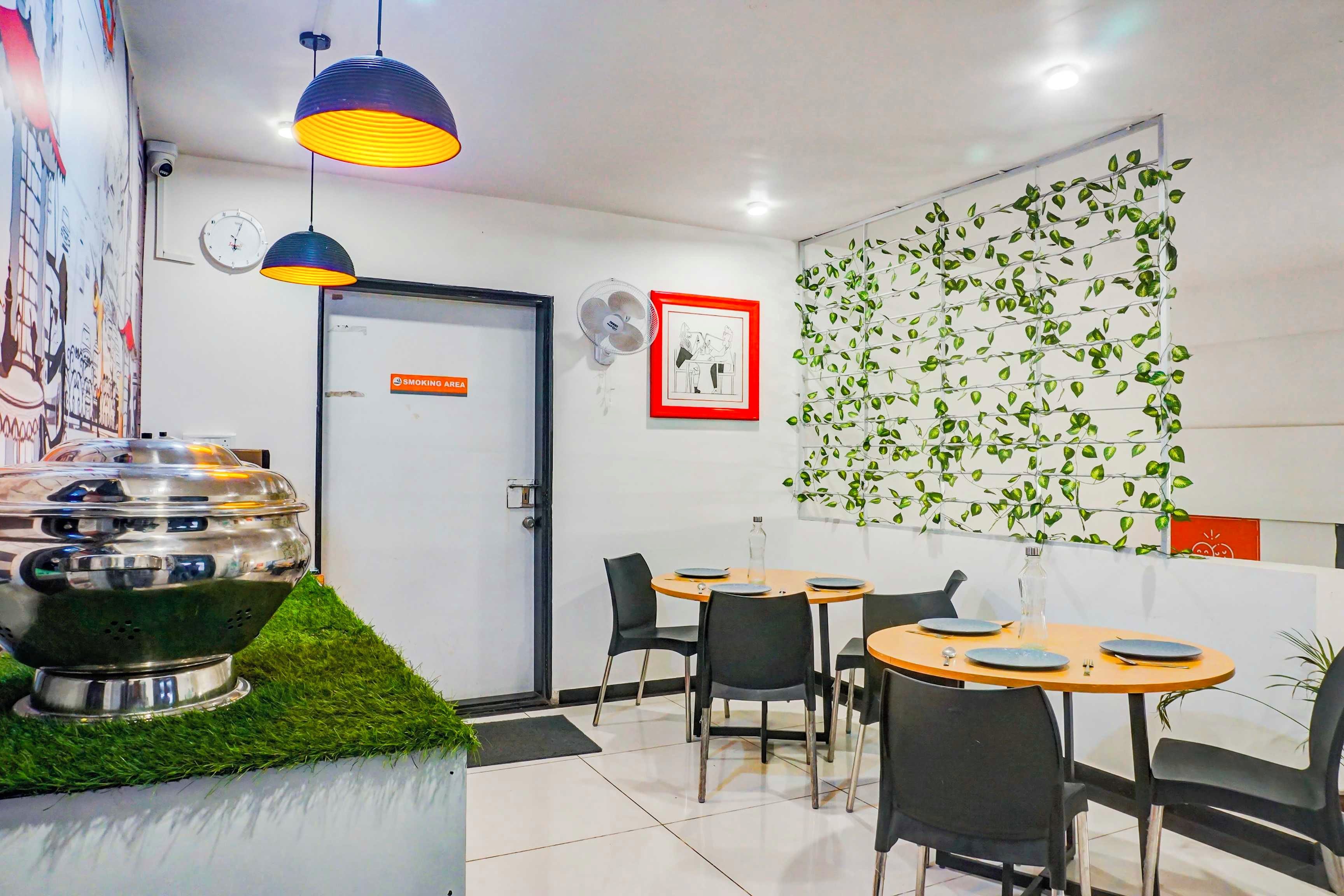 A restaurant interior with round tables, black chairs, a wall-mounted fan, and a decorative wall with green leaves.