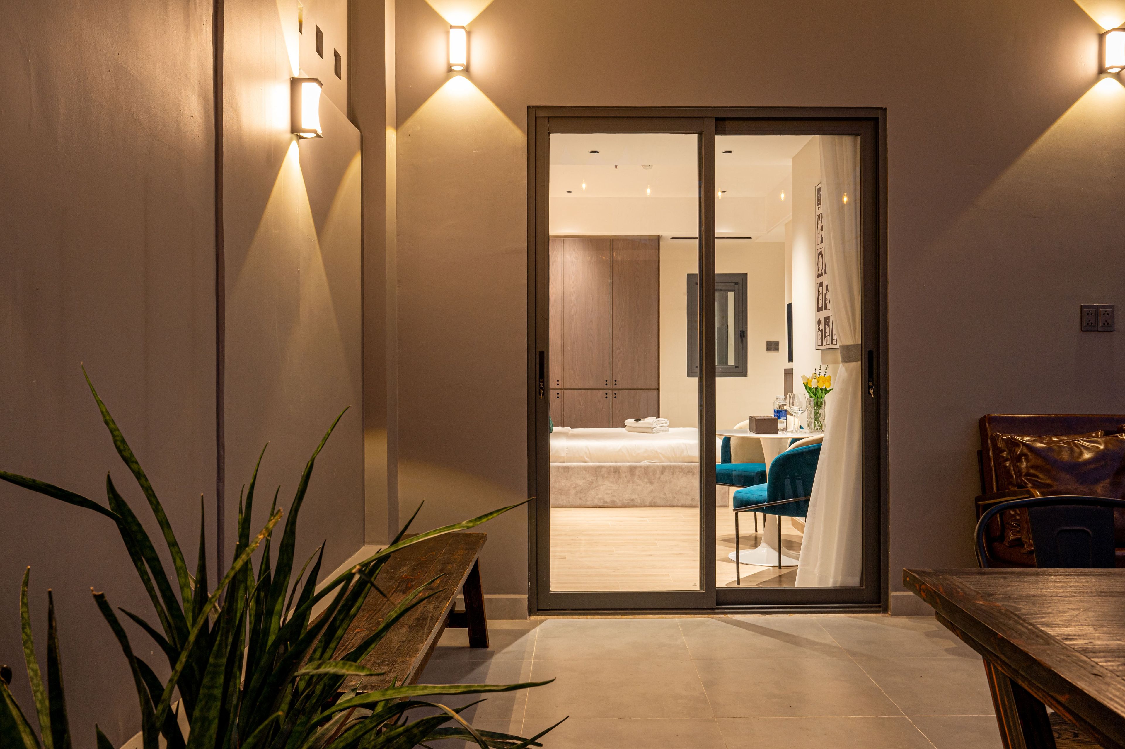 A hotel room with a sliding glass door leading to a bedroom, a dining area with blue chairs, and a wooden table.
