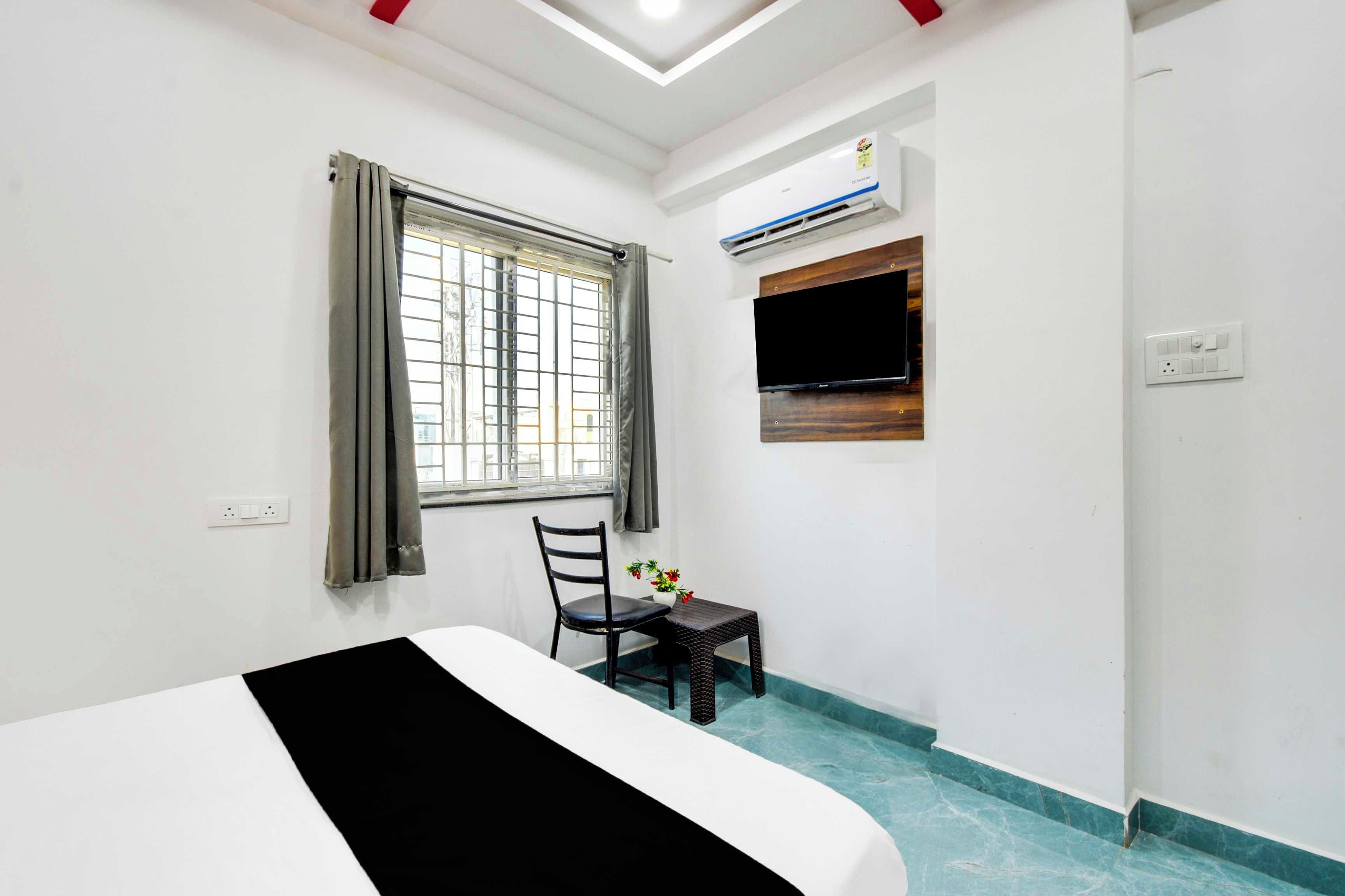 Habitación de hotel con cama, ventana con cortinas, una silla, una mesita con flores y un equipo de aire acondicionado.