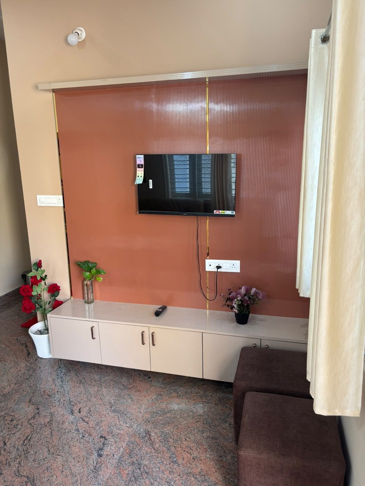 Una habitación con una televisión de pantalla plana montada en la pared, un mueble blanco con un control remoto y un sofá marrón.