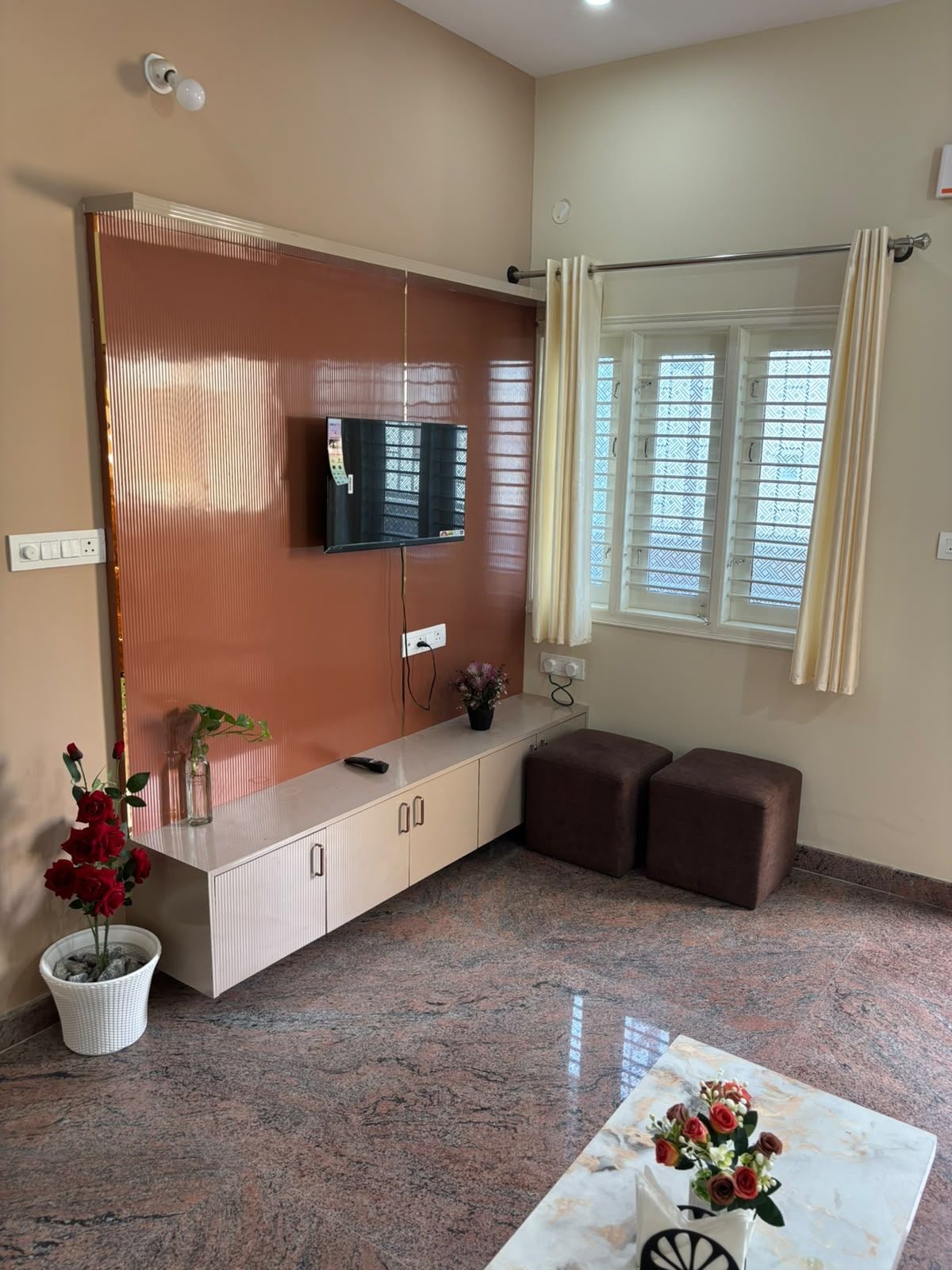 A room with a flat-screen TV mounted on a wall, a window with curtains, a small seating area with two ottomans, and a table with a vase of flowers.