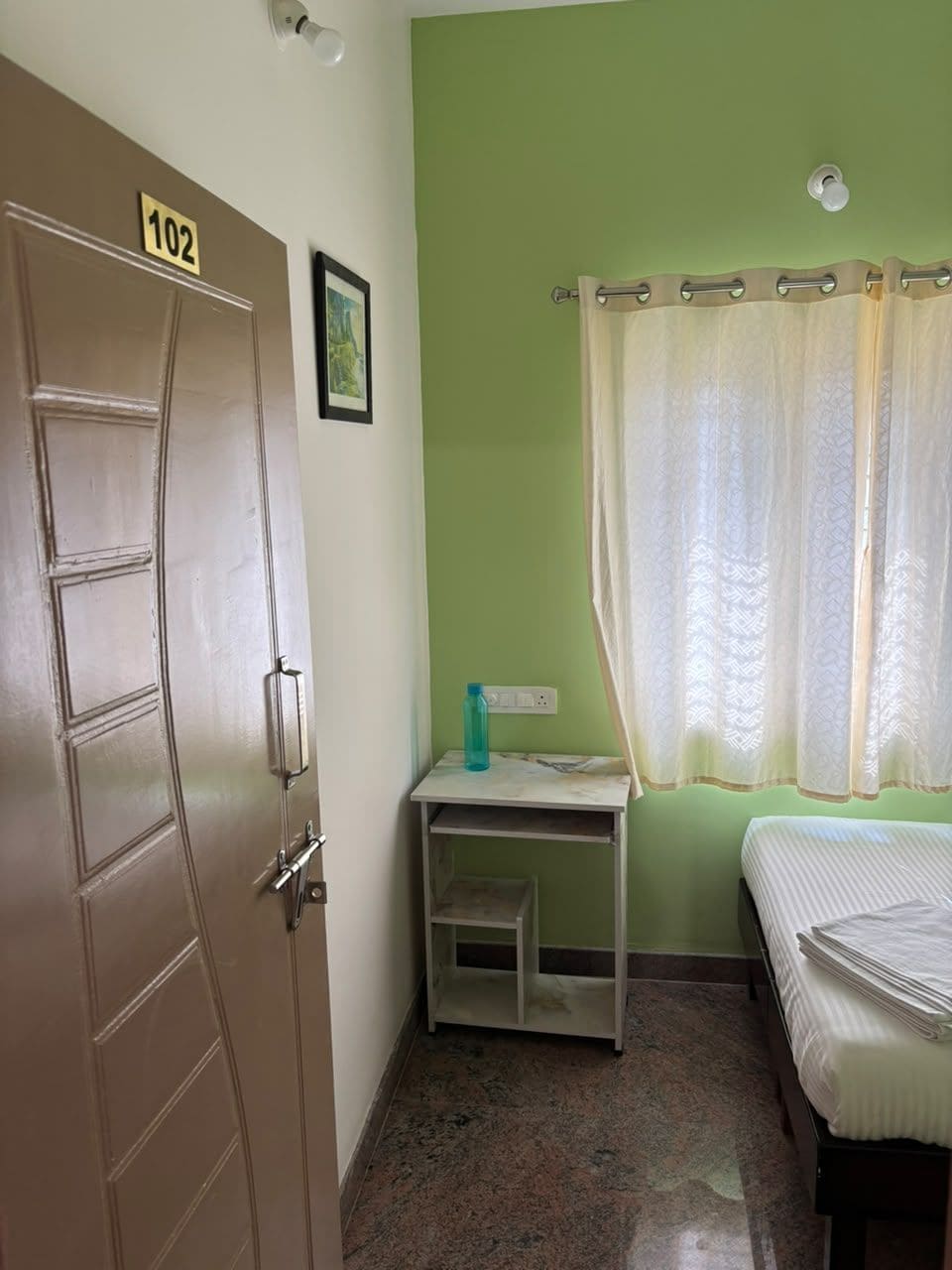Una habitación con una puerta identificada con el número '102', una cama, una mesita con una botella de agua y una ventana con cortina.