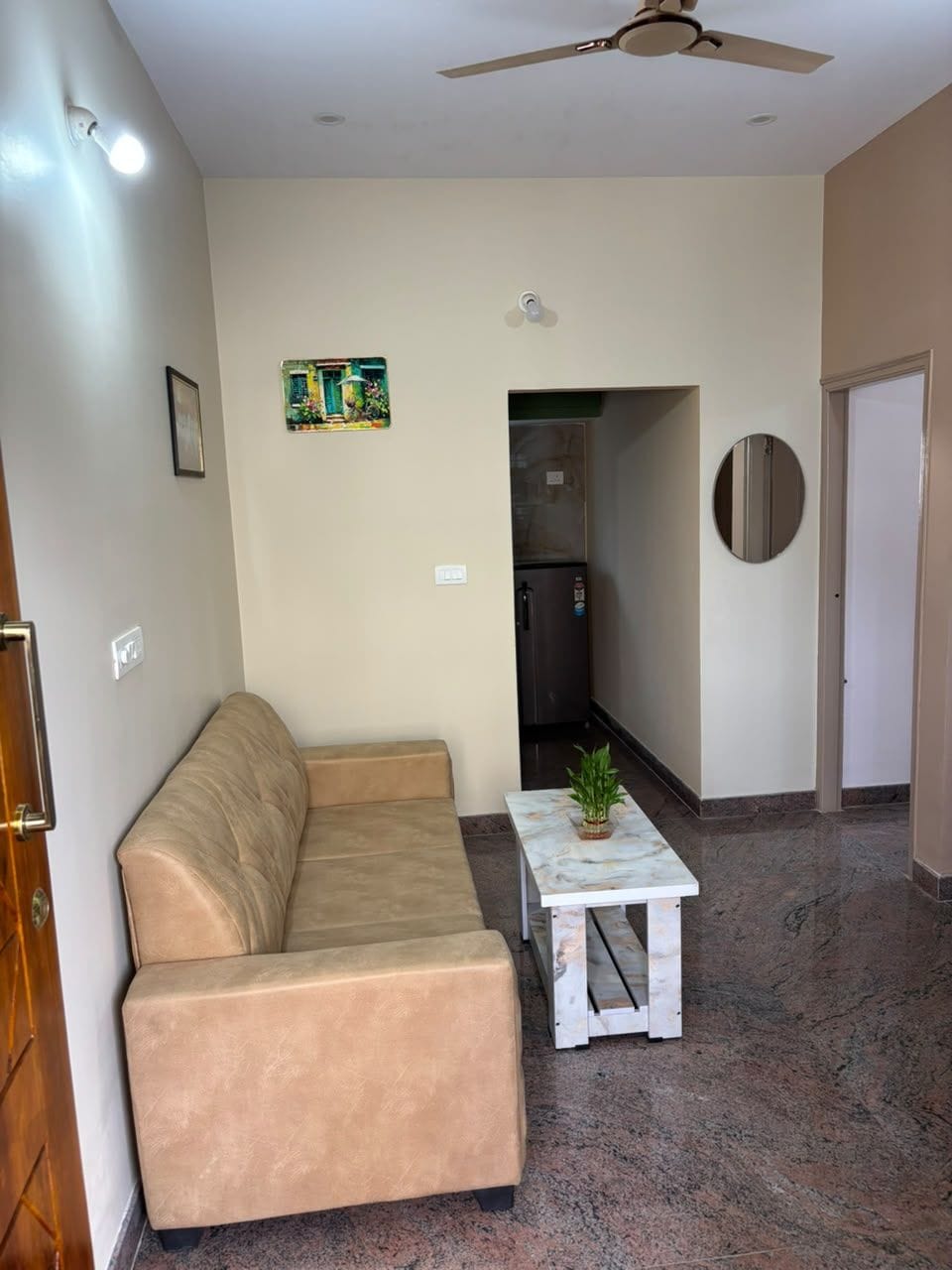 Una sala de estar con un sofá color beige, una mesita con una planta, un ventilador de techo y una cocina al fondo.