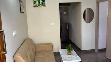Una sala de estar con un sofá color beige, una mesita con una planta, un ventilador de techo y una cocina al fondo.