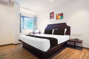 A hotel room with a large bed, a bedside table, a nightstand, a window with curtains, and two wall-mounted artworks.