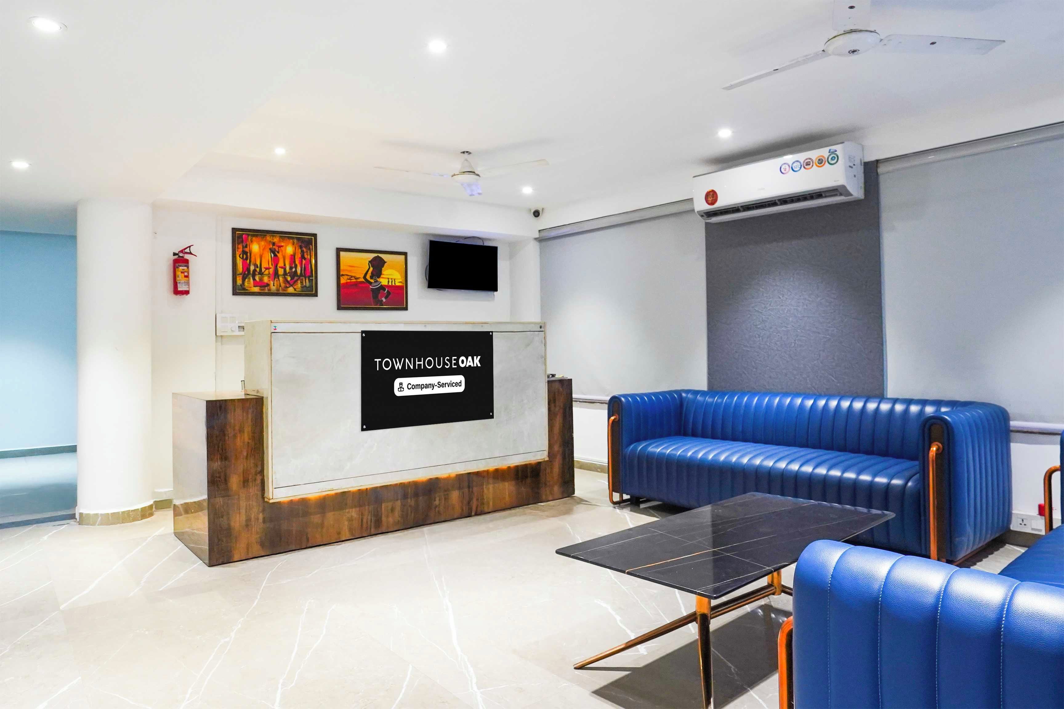 A reception area with a sign reading 'TOWNHOUSE OAK Company-Serviced,' a flat-screen TV, a ceiling fan, and blue leather seating.