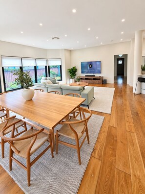 A modern living room with a wooden dining table, comfortable seating, a flat-screen TV, and a view of the outdoors.