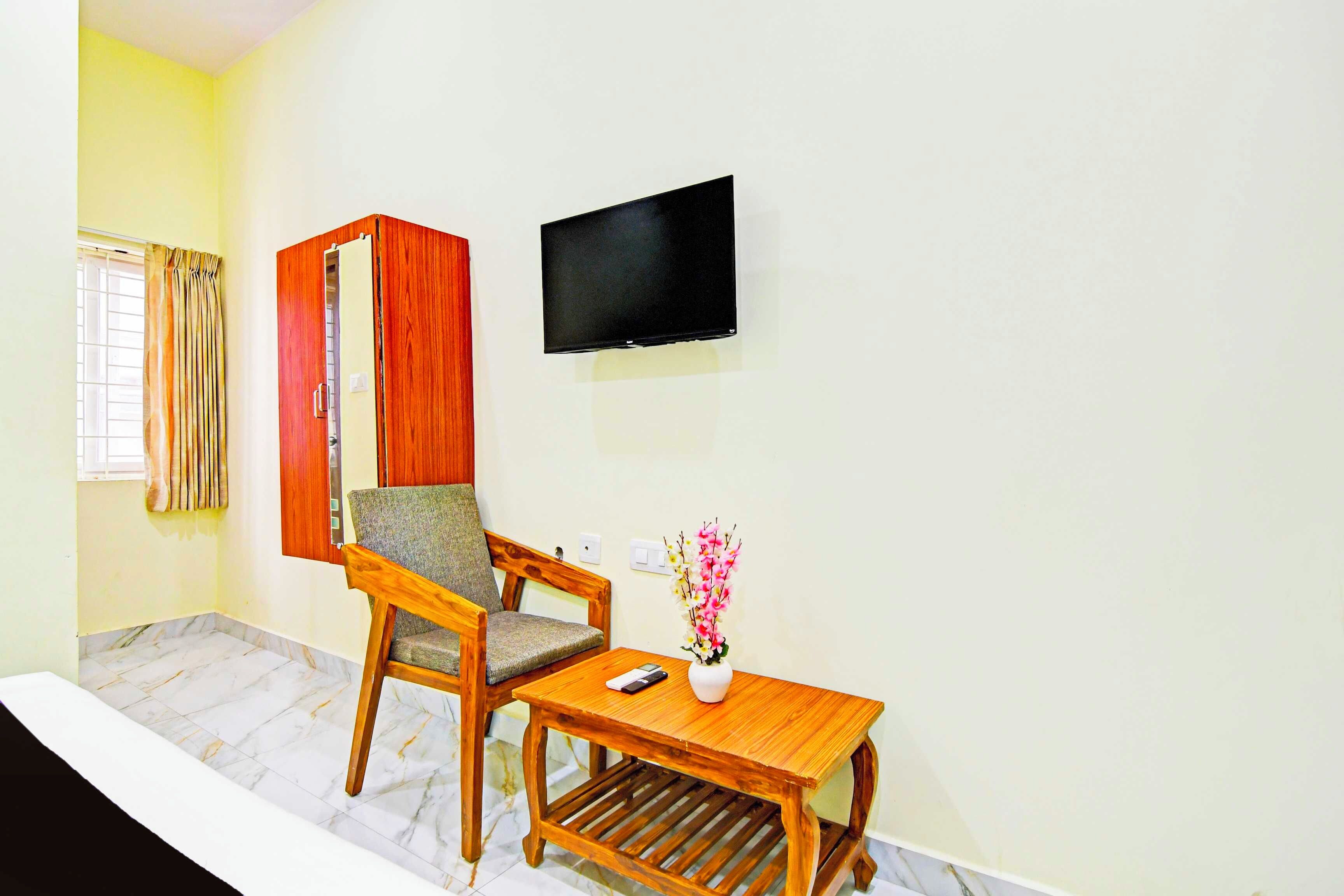 Una habitación con una silla de madera, una pequeña mesa de madera, un televisor de pantalla plana montado en la pared y una ventana con cortinas.