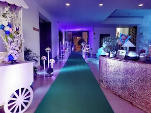 A hotel lobby with a green carpet leading to a reception desk, decorated with flowers and various items.