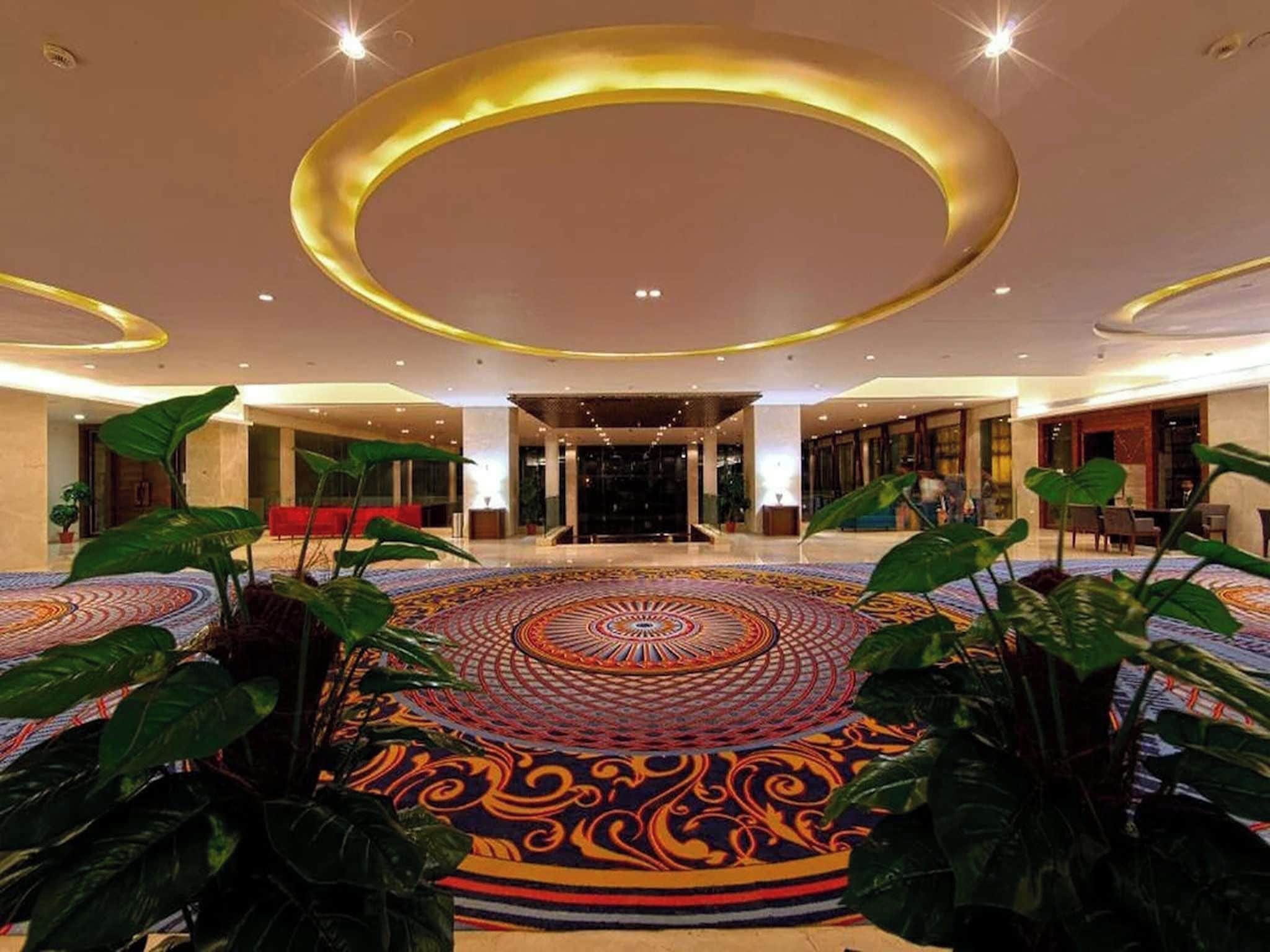 A spacious lobby with a large circular patterned carpet, a central seating area, and potted plants.