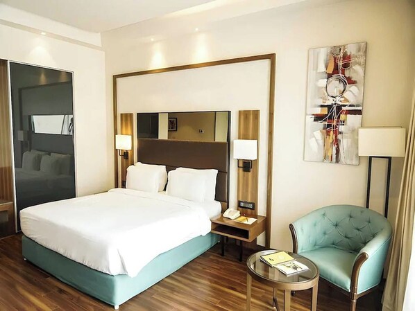 A hotel room with a large bed, bedside tables, a chair, a small table, and a painting on the wall.